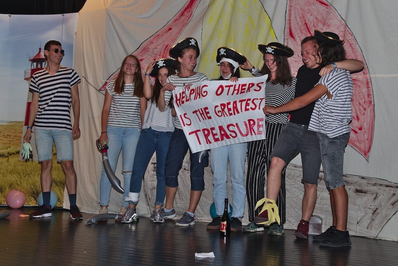 A youth group during their pirate-themed stage play, holding a sign saying "helping others is the greatest treasure".