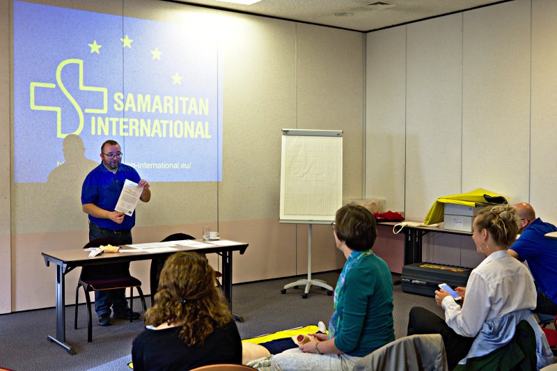 Scene from one of the first aid courses - One of the trainers stands in front of a presentation screen, several participants are listening.