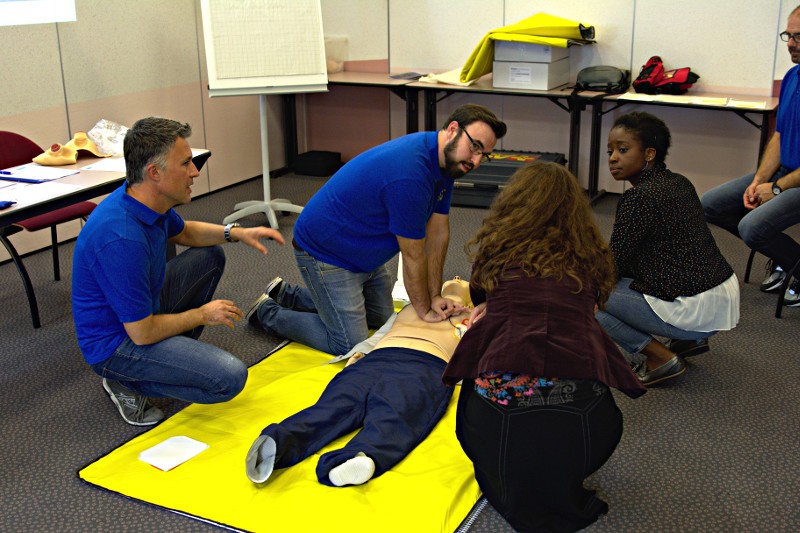 Scene from a course - Two trainers and two participants are next to a CPR mannequin, the two trainers explain and demonstrate how to perform CPR.